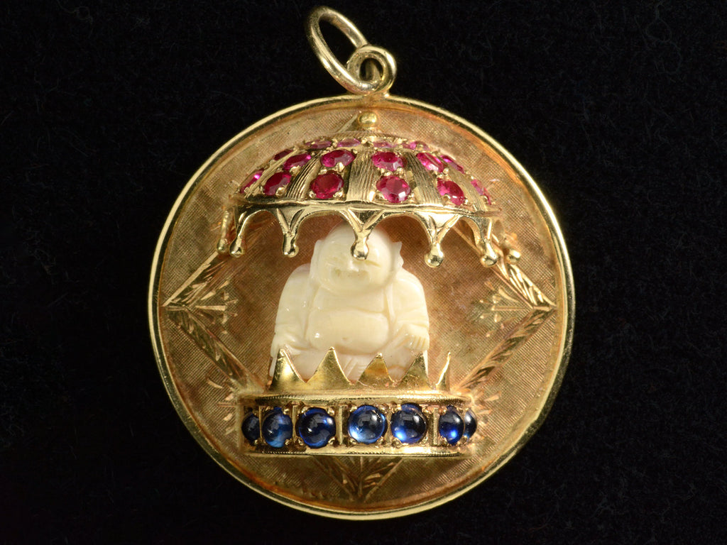 A round  yellow gold vintage pendant with a carved bone Buddha figure beneath a parasol of rubies and resting on a sapphire lined dais. Shown on black background.
