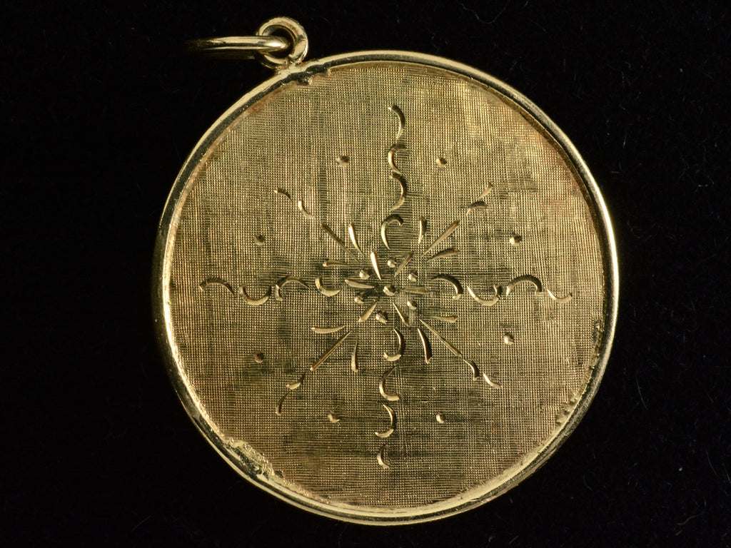 Back of a round  yellow gold vintage pendant with textured surface and engraved designs. Shown on black background.