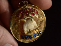 thumbnail of Angled view of a round  yellow gold vintage pendant with a carved bone Buddha figure beneath a parasol of rubies and resting on a sapphire lined dais. Shown held in fingers.