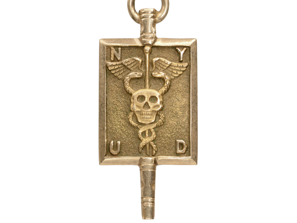 A rectangular pendant with a skull and a caduceus and the letters N, Y, U and D. In yellow gold. On white background.