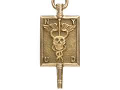 thumbnail of A rectangular pendant with a skull and a caduceus and the letters N, Y, U and D. In yellow gold. On white background.