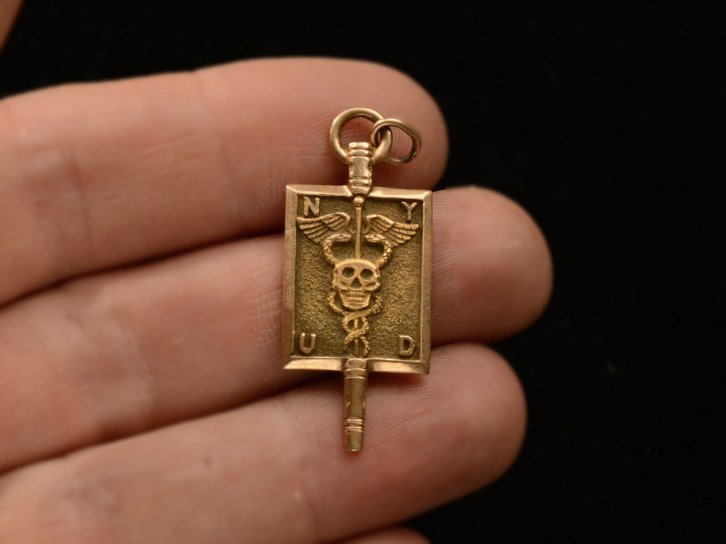 A rectangular pendant with a skull and a caduceus and the letters N, Y, U and D. In yellow gold. On hand for scale.