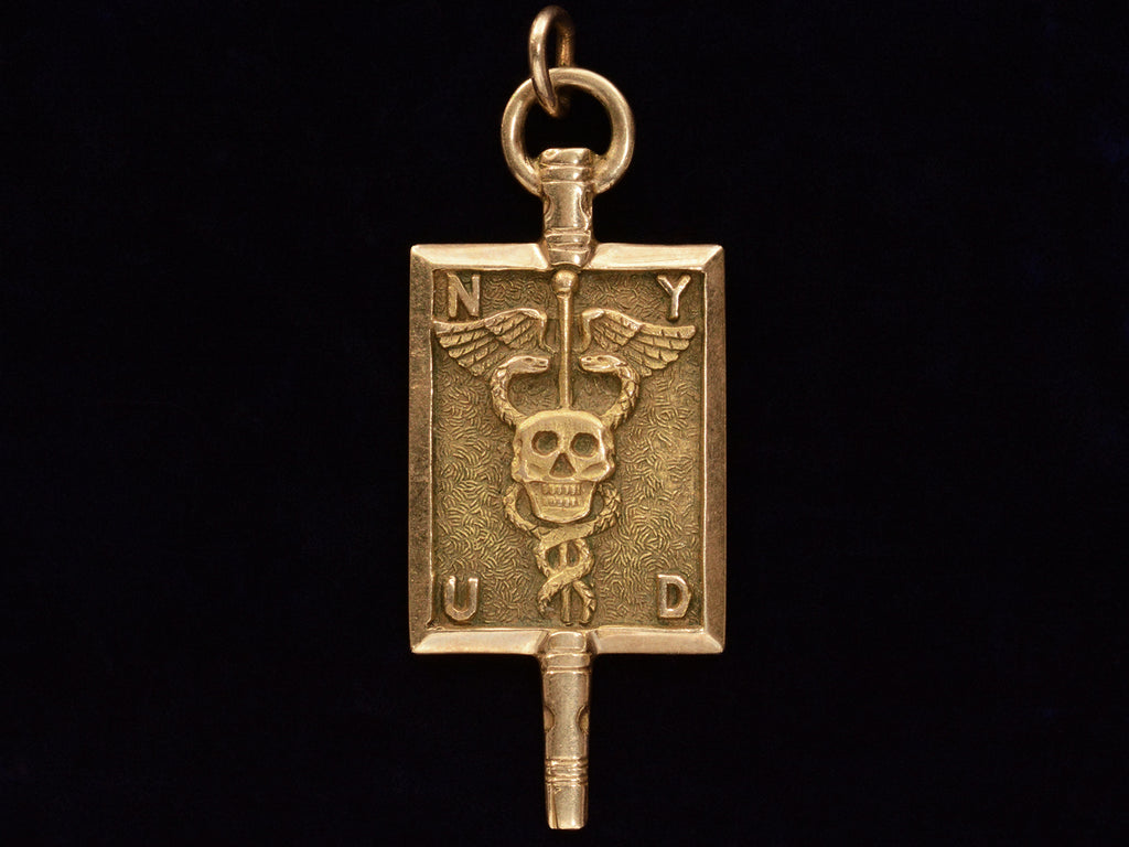 A rectangular pendant with a skull and a caduceus and the letters N, Y, U and D. In yellow gold. On black background.