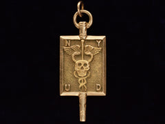 thumbnail of A rectangular pendant with a skull and a caduceus and the letters N, Y, U and D. In yellow gold. On black background.