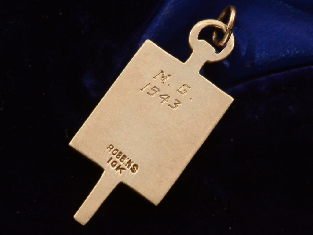 Reverse side view of rectangular pendant with an inscriptions reading "M.G. 1943)" and a stamp that reads "ROBBINS 10K."  In yellow gold. On dark blue background.