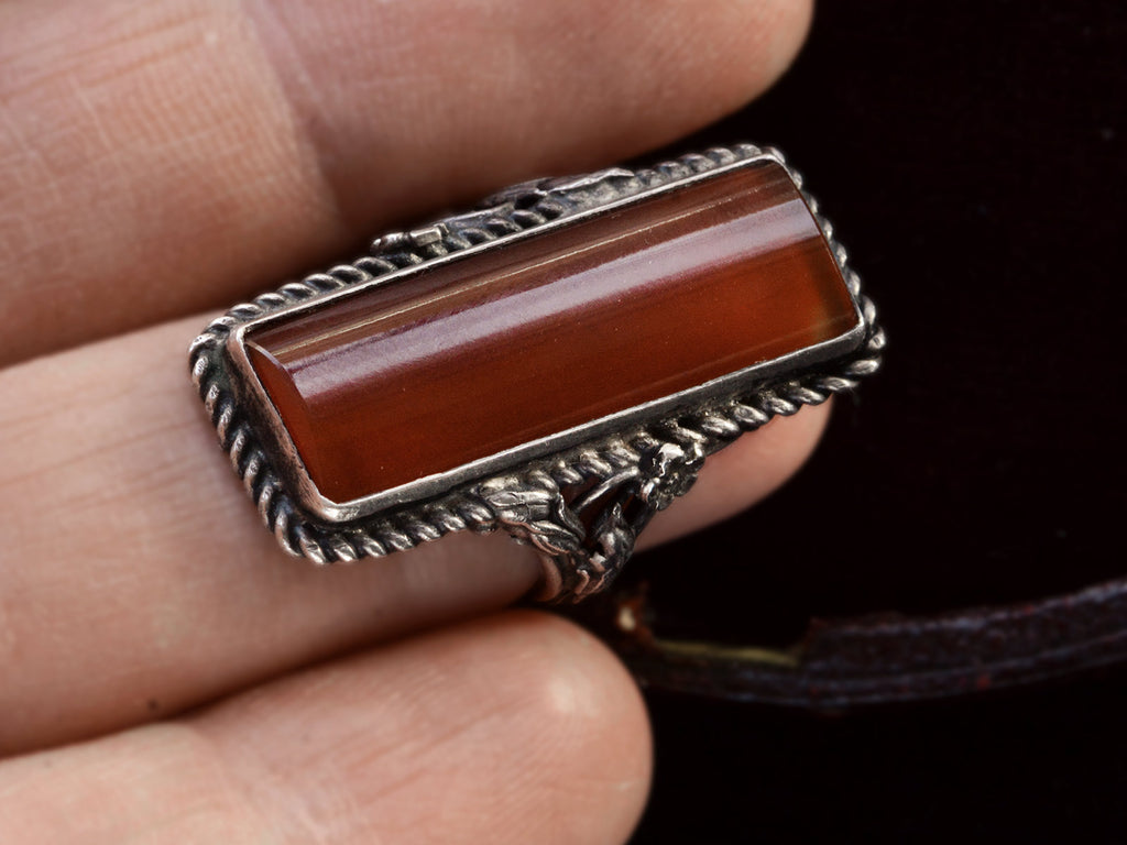 A vintage silver ring with intricate detailing and a long rectangular carnelian stone, shown on finger.