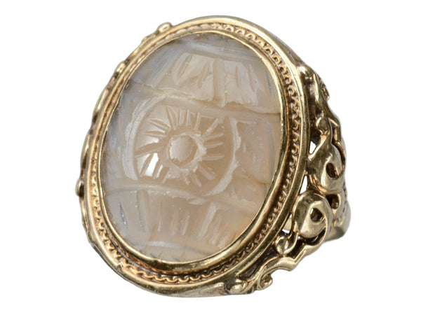 Angled front view of c1920 Art Deco ring with an oval carved blueish brown chalcedony set in a finely detailed gold mounting, shown on white background.