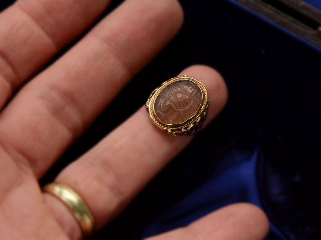 Front view of c1920 Art Deco ring with an oval carved blueish brown chalcedony set in a finely detailed gold mounting, shown on finger