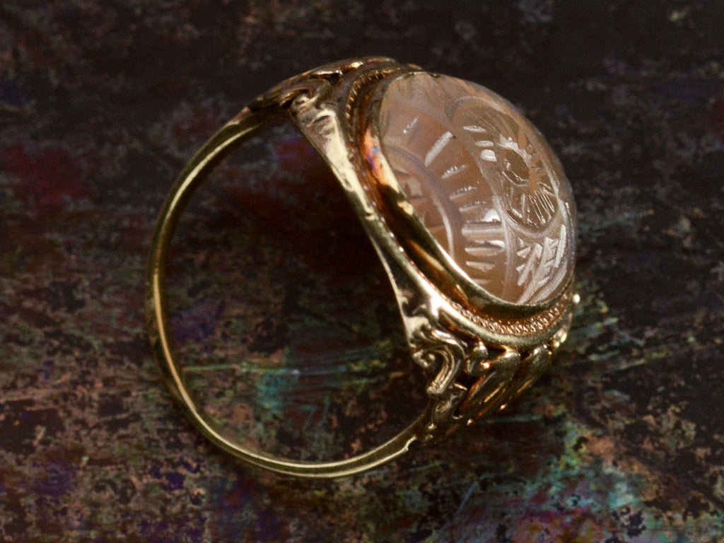 Side profile view of c1920 Art Deco ring with an oval carved blueish brown chalcedony set in a finely detailed gold mounting, shown on dark background.