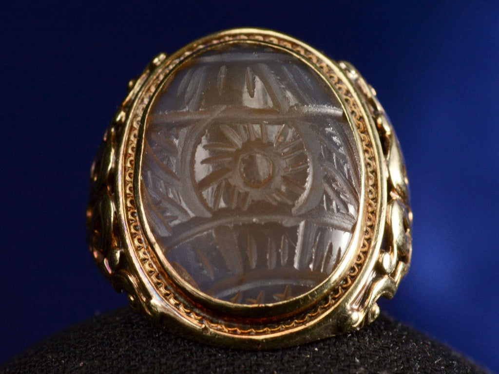 Front view of c1920 Art Deco ring with an oval carved blueish brown chalcedony set in a finely detailed gold mounting, shown on blue background.