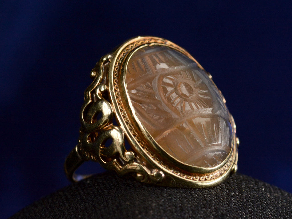 Angled view of c1920 Art Deco ring with an oval carved blueish brown chalcedony set in a finely detailed gold mounting, shown on dark background.