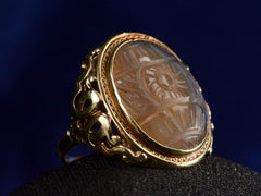 thumbnail of Angled view of c1920 Art Deco ring with an oval carved blueish brown chalcedony set in a finely detailed gold mounting, shown on dark background.