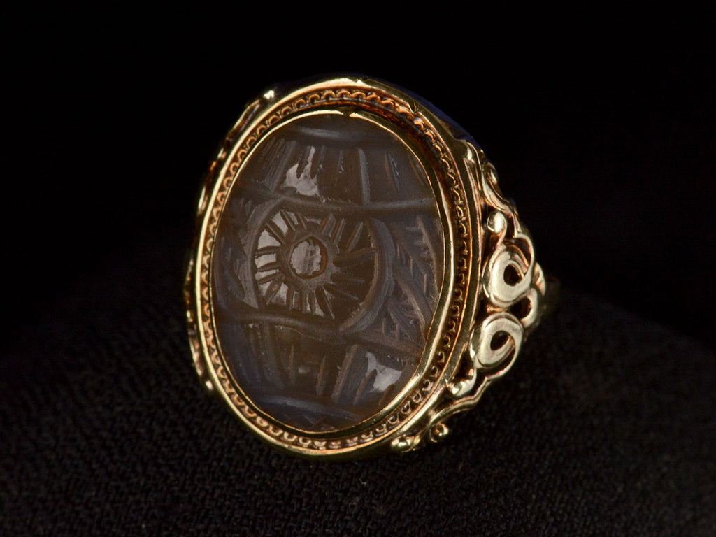 Elevated angled front view of c1920 Art Deco ring with an oval carved blueish brown chalcedony set in a finely detailed gold mounting, shown on dark background.