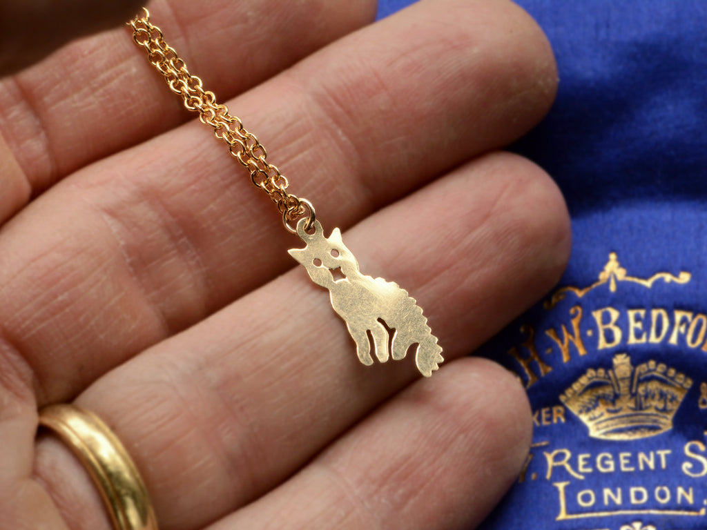 A 2-dimensional yellow gold cat charm hanging on chain, shown on hand.