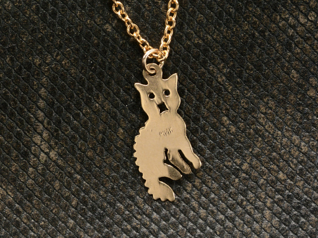 Reverse side of 2-dimensional yellow gold cat charm hanging on chain, shown on dark textured background. "14K" stamp visible.