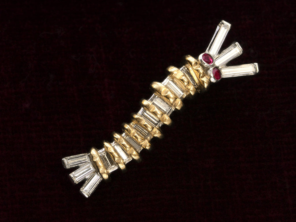 A vintage white and yellow gold caterpillar brooch with ruby eyes and diamond baguettes, on dark background.