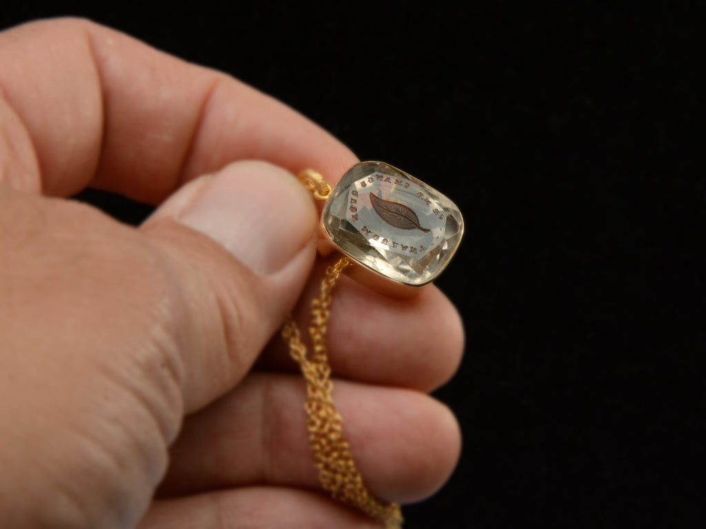 A c1820 Georgian era gold and crystal intaglio pendant carved with the image of an ivy leaf and the French phrase "Je Ne Change Qu'en Mourant." Shown in hand.