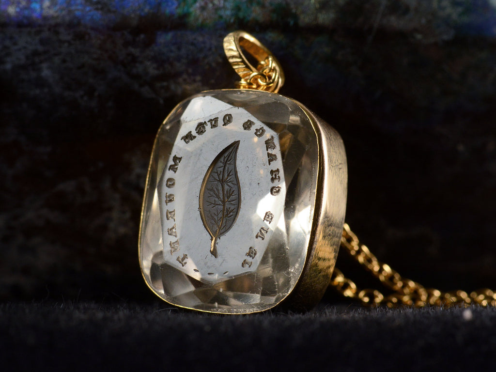 Angled detail view of c1820 Georgian era gold and crystal intaglio pendant carved with the image of an ivy leaf and the French phrase "Je Ne Change Qu'en Mourant." Shown on dark background.