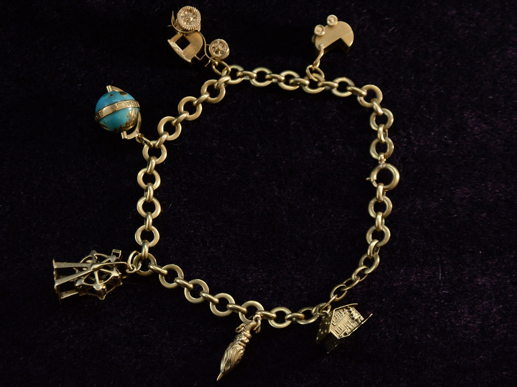 A vintage c1940 yellow gold charm bracelet with charms depicting a house, an umbrella, a ferris wheel, a blue enamel globe, a car, and a baby carriage. Shown on dark background.