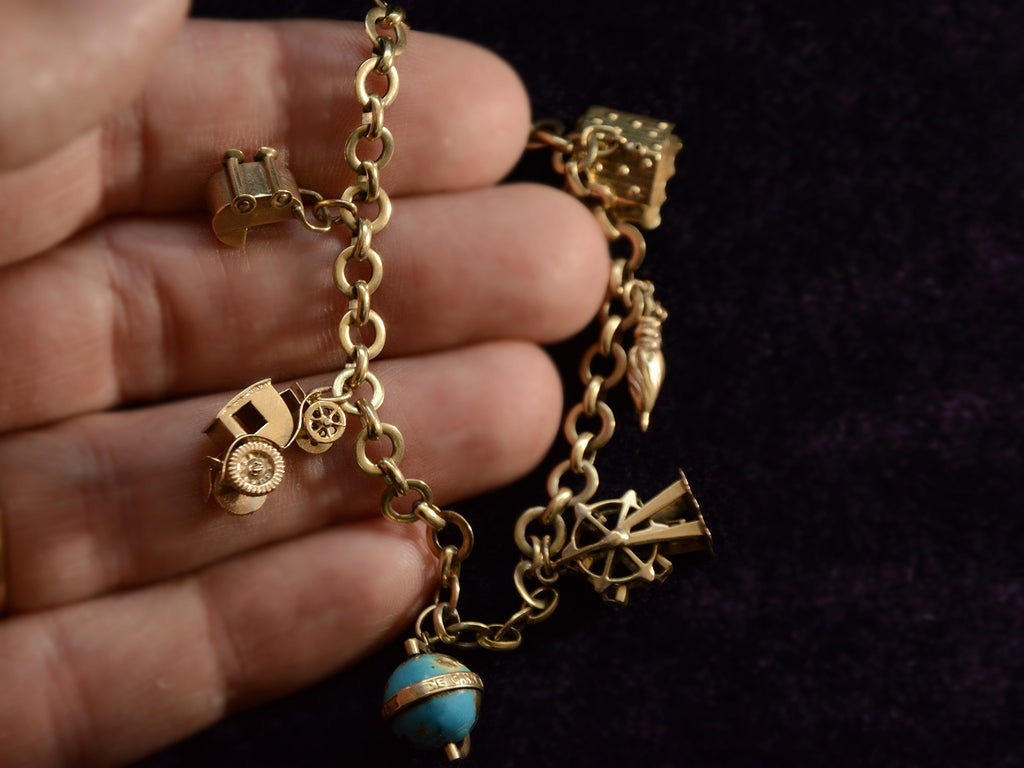 Hand holding a vintage c1940 yellow gold charm bracelet with charms depicting a house, an umbrella, a ferris wheel, a blue enamel globe, a car, and a baby carriage. Shown on dark background.