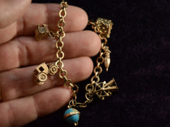 thumbnail of Hand holding a vintage c1940 yellow gold charm bracelet with charms depicting a house, an umbrella, a ferris wheel, a blue enamel globe, a car, and a baby carriage. Shown on dark background.