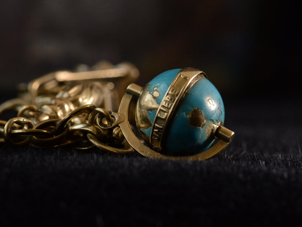 Detail view of yellow gold and blue enamel globe charm with the German words "UM LIEBE" visible. On dark background.