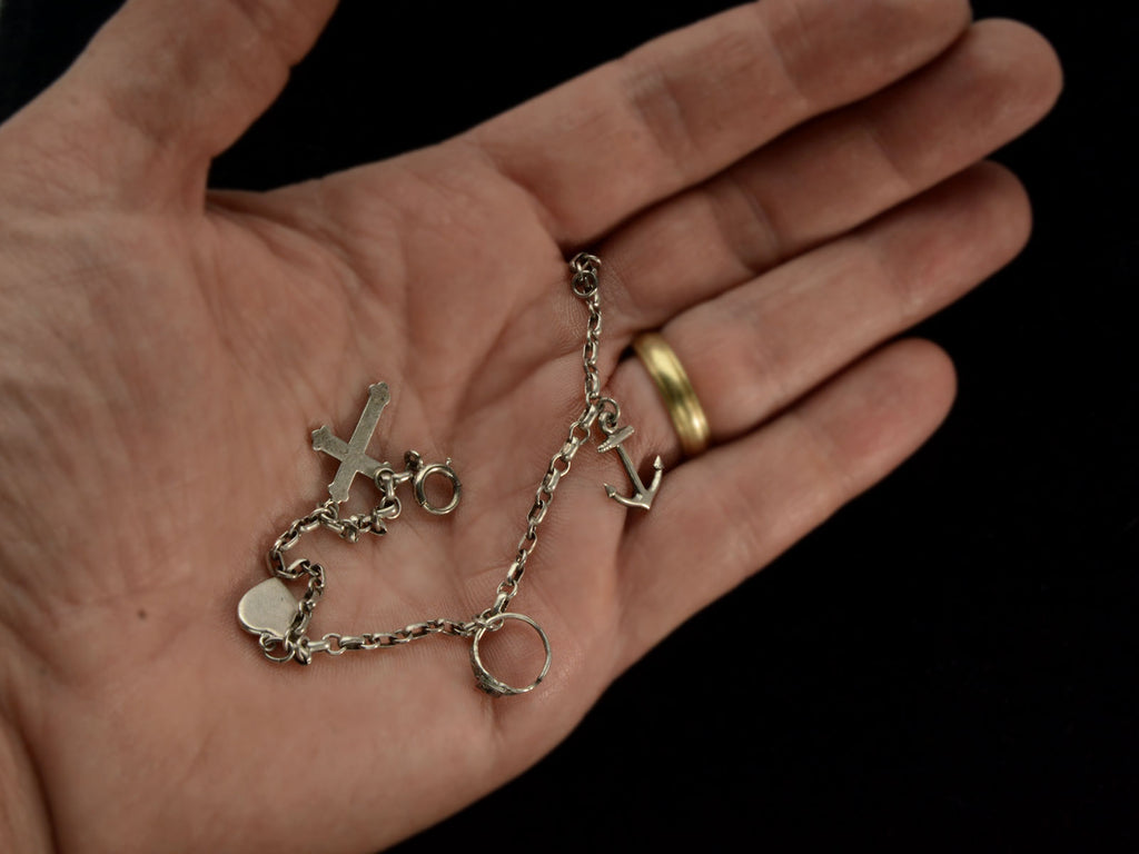 Hand holding a c1930 Vintage Silver Charm Bracelet with 4 Charms: an Anchor, a Baby Ring, a Heart, and a Cross. 
