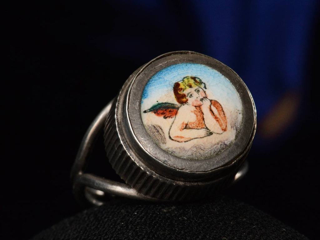 Left angle view of c1900 Silver and Enamel Cherub Ring (on dark blue background)