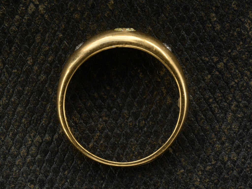Side profile of a rounded vintage 18k yellow gold ring flush set with a light greenish-yellow round diamond in between two smaller light pink diamonds, shown dark textured background.