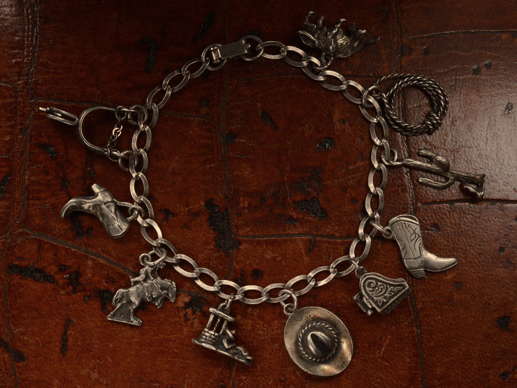 Vintage western-themed silver charm bracelet with a spur, two boots, a horse, a man sleeping in a sombrero, a cowboy hat, a piano with functioning lid, a cactus, a rope and a bull, on dark leather background.