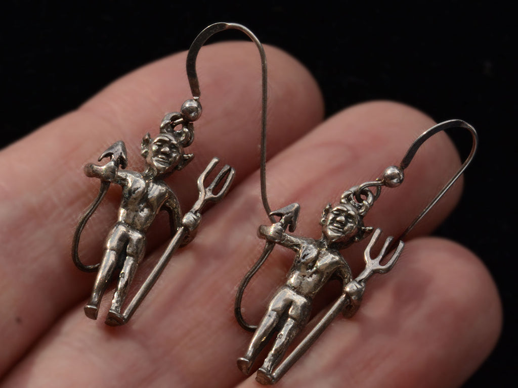 Pair of silver devil earrings with pitchforks, shown on hand.