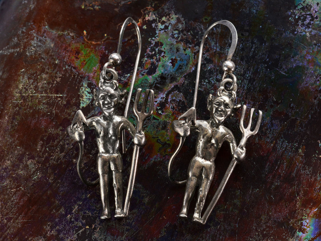 Pair of silver devil earrings with pitchforks, shown on dark textured background.