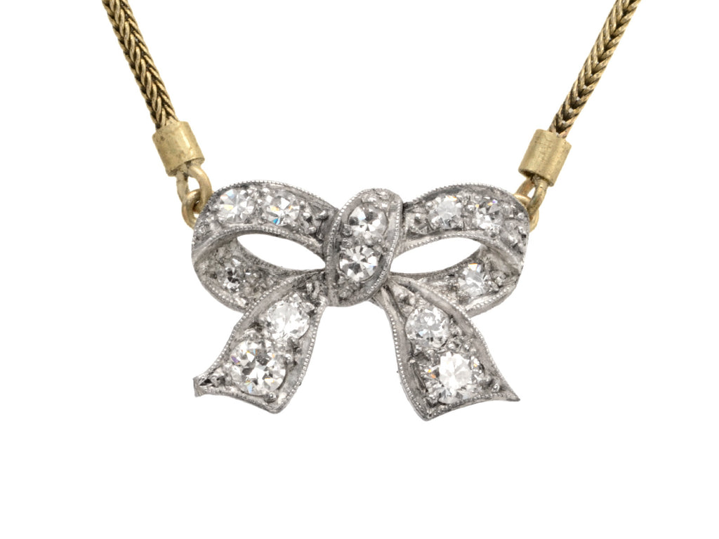 Detail of an antique Edwardian-era necklace with a silver bow-shaped pendant set with diamonds, hanging from a yellow gold snake chain, shown on white background