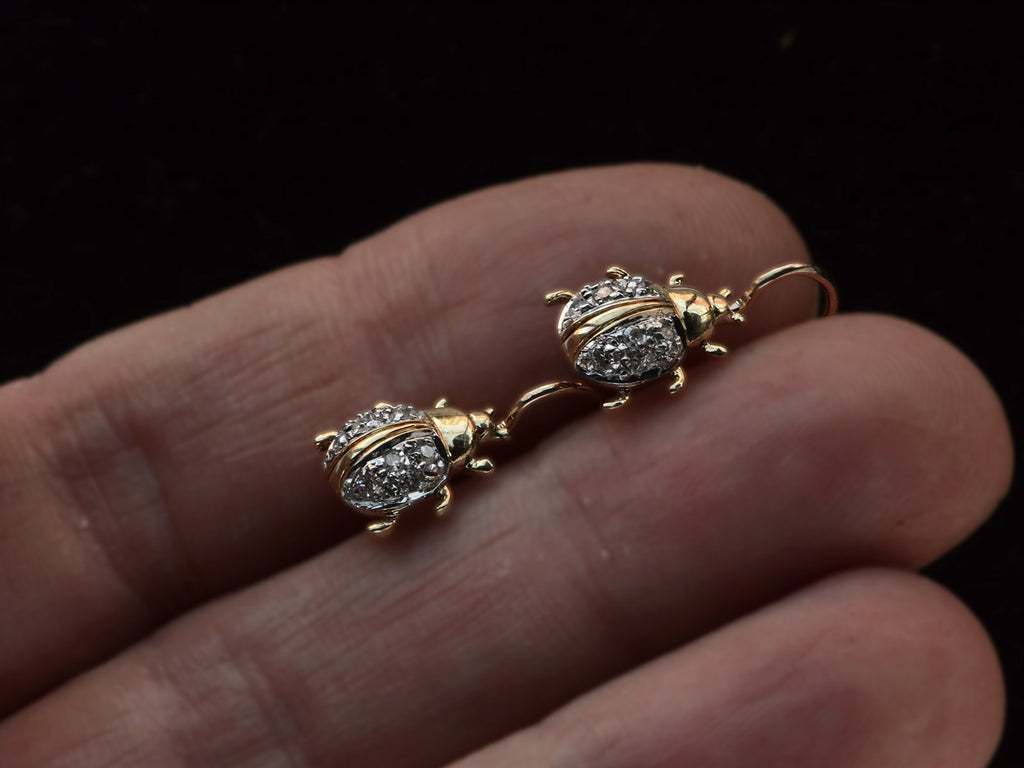 A pair of vintage bug-shaped earrings in yellow gold with white gold details and single cut diamonds set in the bodies, held in between fingers.