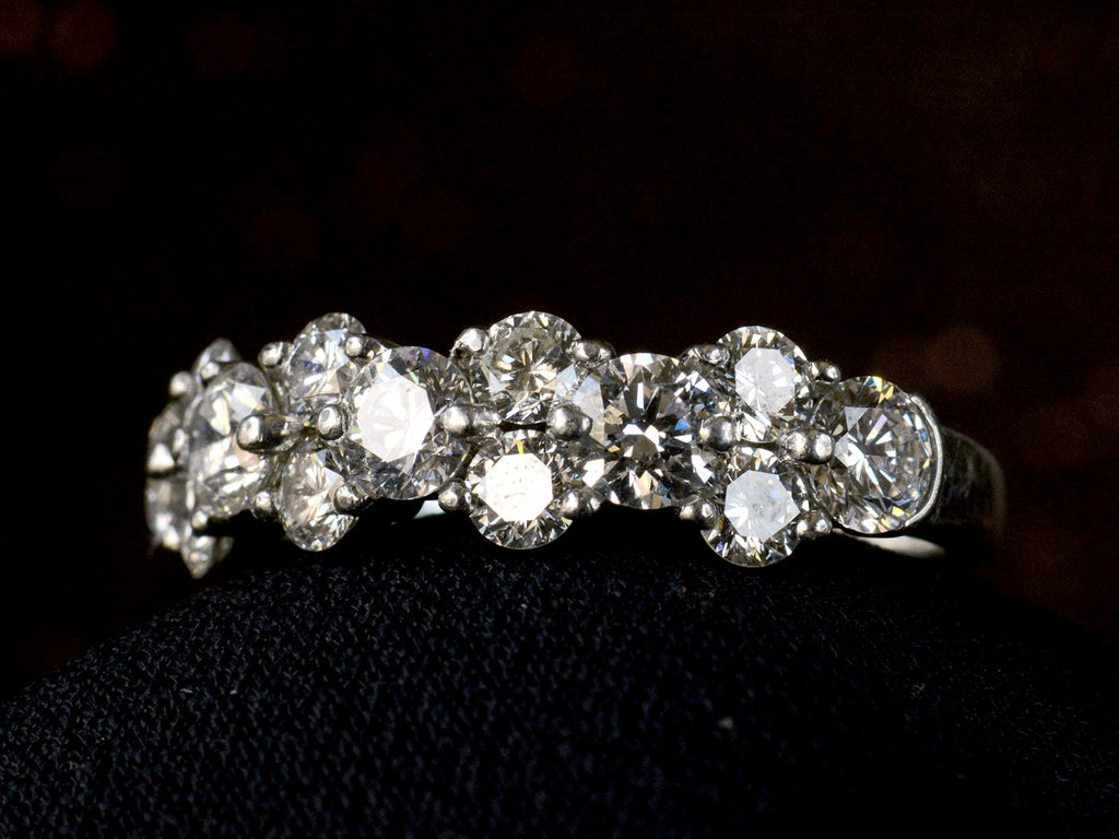 Right angled view of band like platinum ring, half encrusted with a cluster of round diamonds, shown on dark background. 