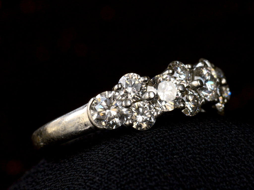 Left angled view of band like platinum ring, half encrusted with a cluster of round diamonds, shown on dark background. 
