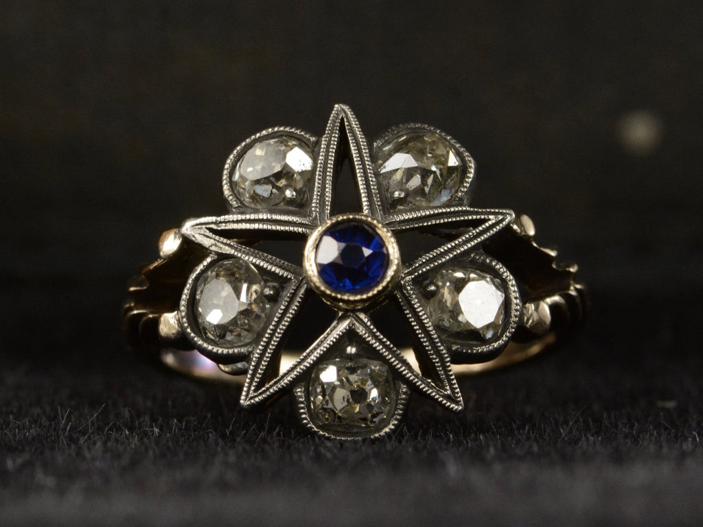 Front view of an antique silver-topped rose gold Victorian ring with a star design centering on a blue sapphire and old mine cut diamonds set in between each point. Shown on dark background.