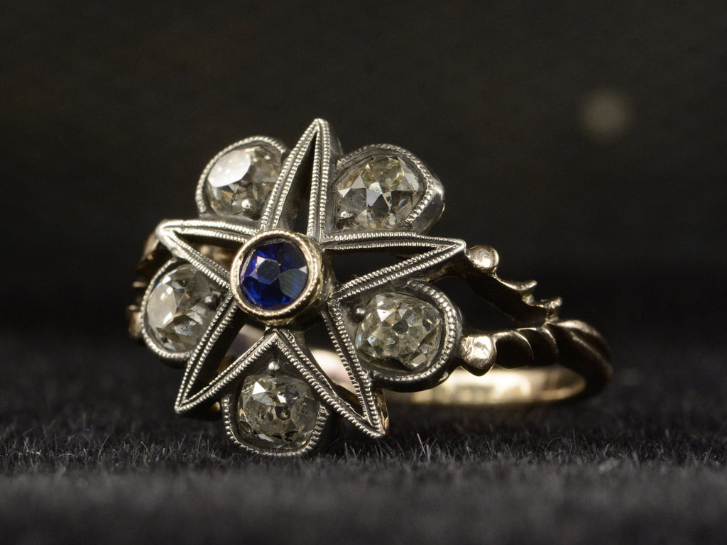 Angled view of an antique silver-topped rose gold Victorian ring with a star design centering on a blue sapphire and old mine cut diamonds set in between each point. Shown on dark background.