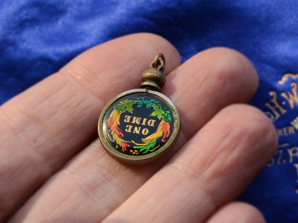 Opposite side of pendant with gilt and colorful enamel dime mounted in brass, shown on hand. The words "One Dime" are visible on the dime.