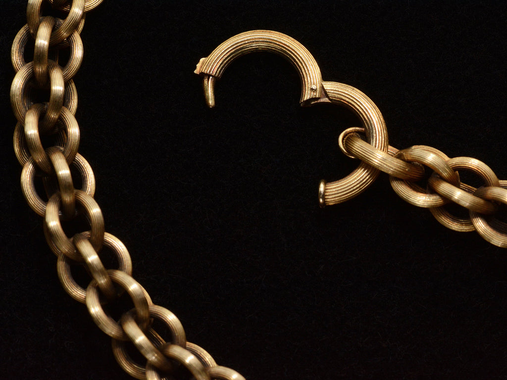 Detail view of a double-link chain necklace with reeded texture, showing the clasp open. On dark background.