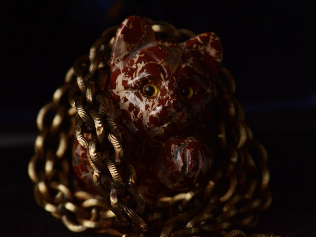 A double-link chain necklace with reeded texture, shown wrapped around a small carved red stone cat figure. Detail view shows one link with small dent.