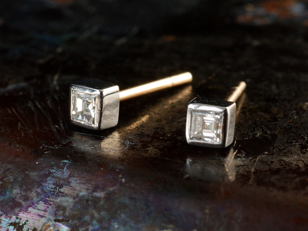 Angled front view of a pair of 0.19cttw square step cut white diamond stud earrings in platinum bezels with yellow gold posts, shown on dark textured background.