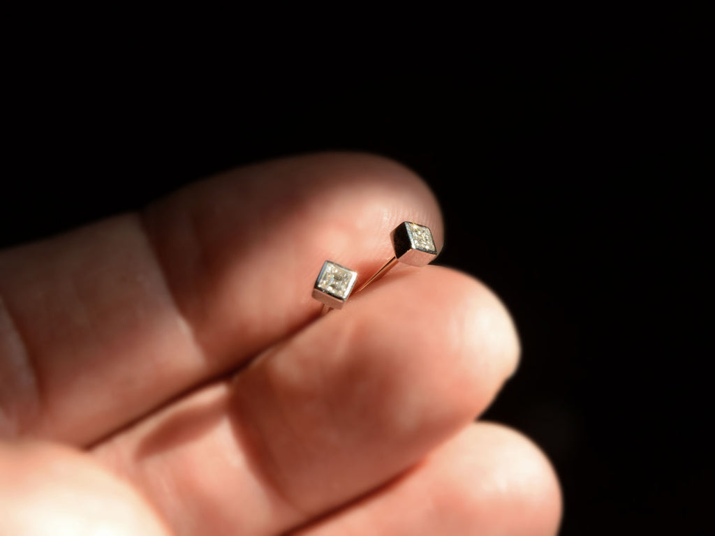 Pair of 0.19cttw square step cut white diamond stud earrings in platinum bezels with yellow gold posts, shown on hand for scale.