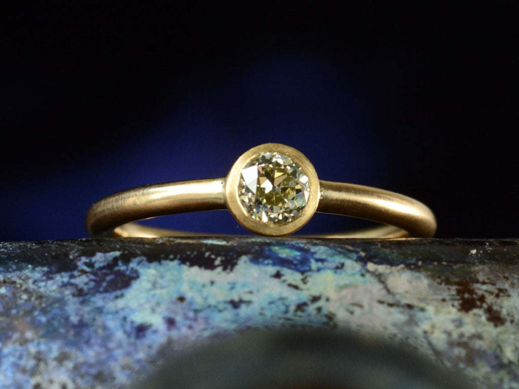 Front view of simple EB Modern ring with a round light yellow diamond in a yellow gold bezel. Shown dark colorful background.