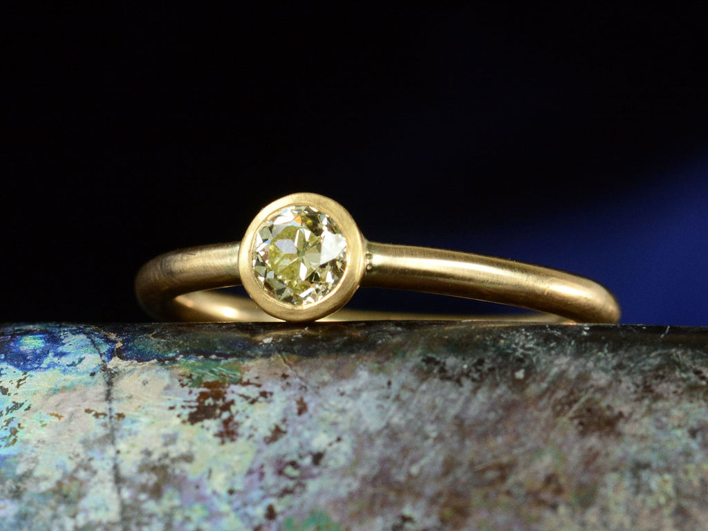 Right angled front view of simple EB Modern ring with a round light yellow diamond in a yellow gold bezel. Shown dark colorful background.
