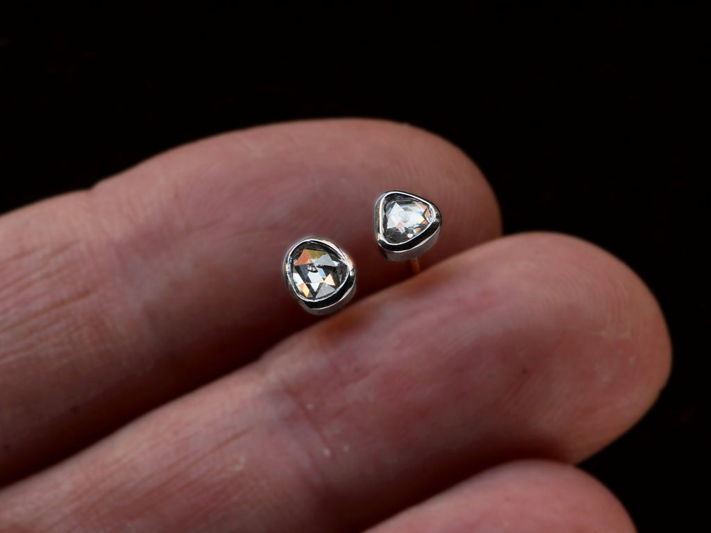 A pair of mismatched and irregular shaped rose cut diamonds set in platinum bezels as stud earrings, shown held between fingers