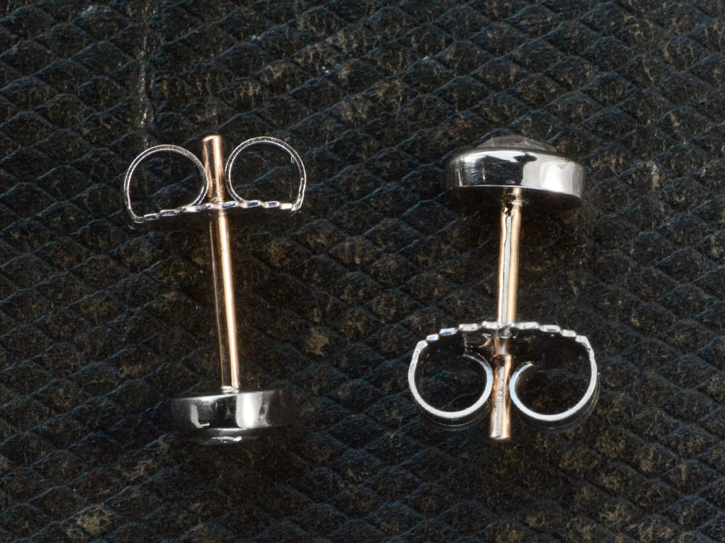 Side profiles of a pair of mismatched and irregular shaped rose cut diamonds set in platinum bezels as stud earrings with 18K yellow gold posts and 18K white gold butterfly backs, shown on dark textured background