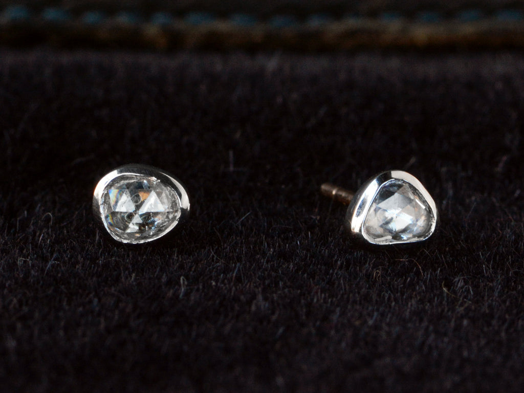 A pair of mismatched and irregular shaped rose cut diamonds set in platinum bezels as stud earrings, shown on dark background