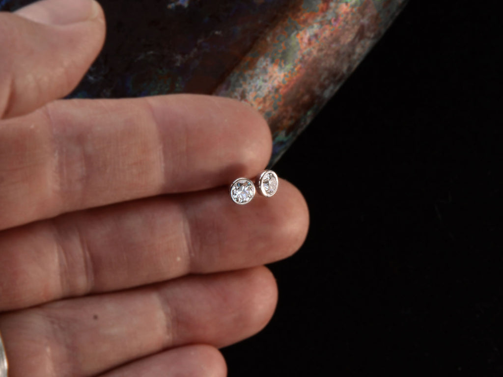 A pair of EB Modern stud earrings with platinum bezels and round white old cut diamonds. Shown held in hand.