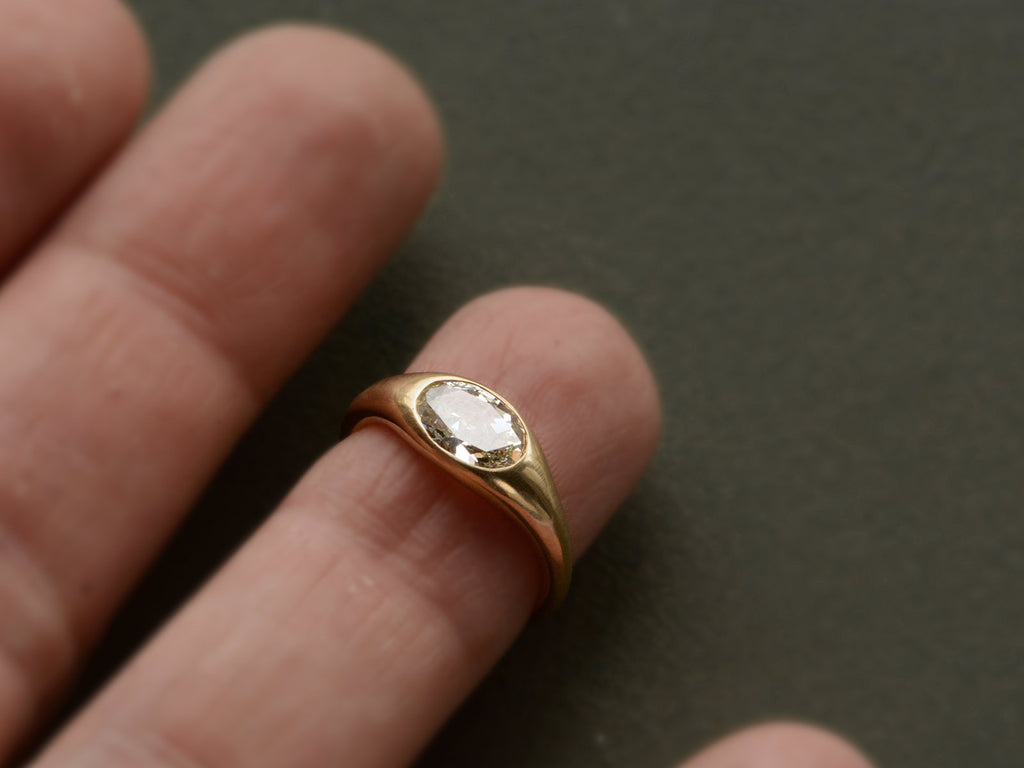 Flush set oval diamond ring in yellow gold, shown on finger against dark green background.